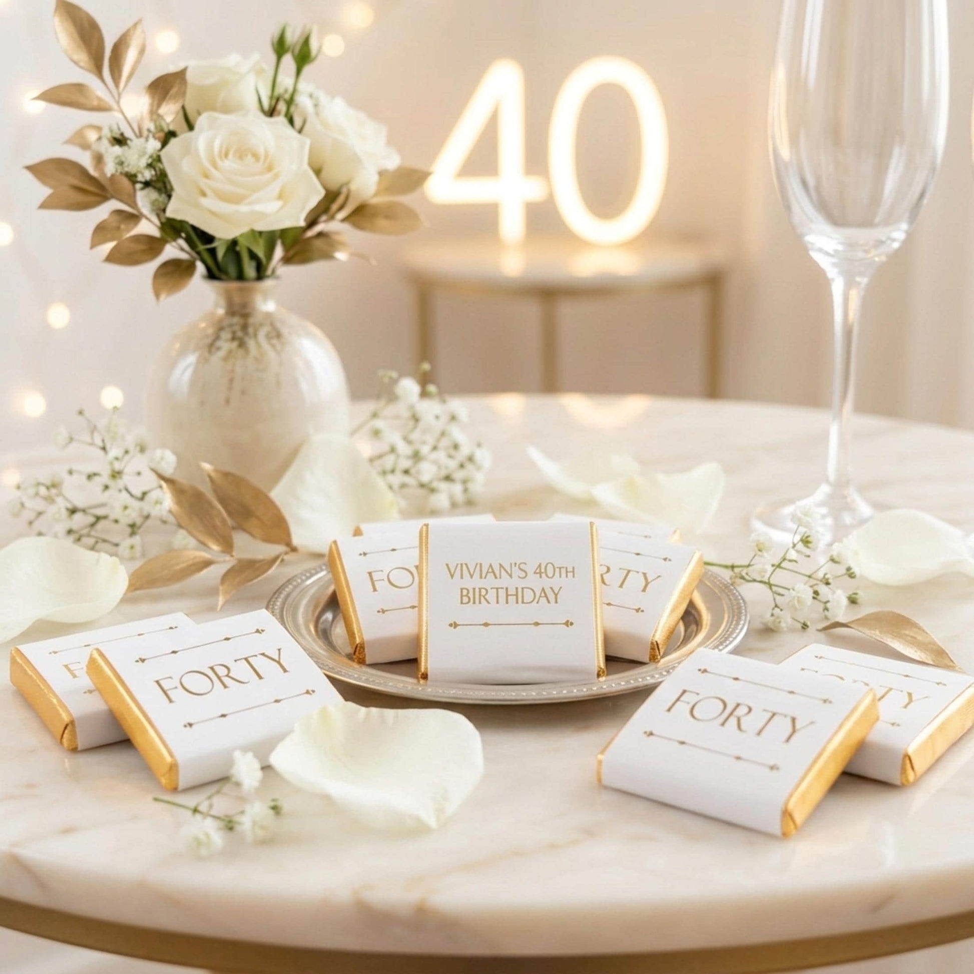 May include: A birthday celebration with white and gold decorations. A vase of white roses and gold leaves is next to chocolates reading "FORTY" and "Vivian's 40th Birthday." A neon "40" sign glows in the background, with a champagne glass.
