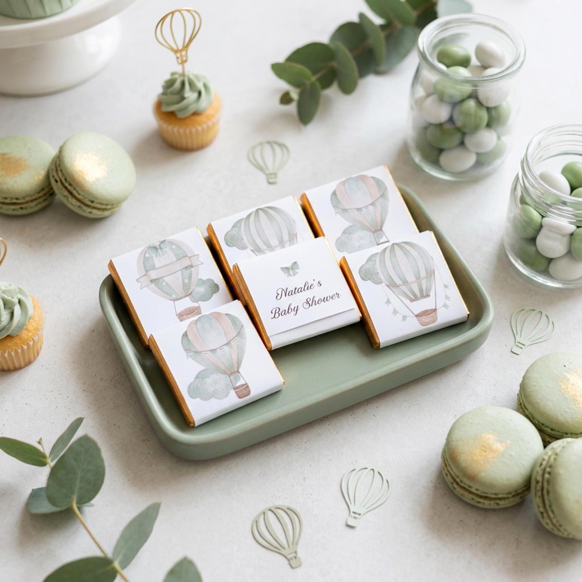 May include: A baby shower arrangement with chocolate bars decorated with hot air balloon designs, presented on a green tray. The text "Natalie's Baby Shower" is printed on one bar. Macarons, cupcakes, and sweets are also visible.