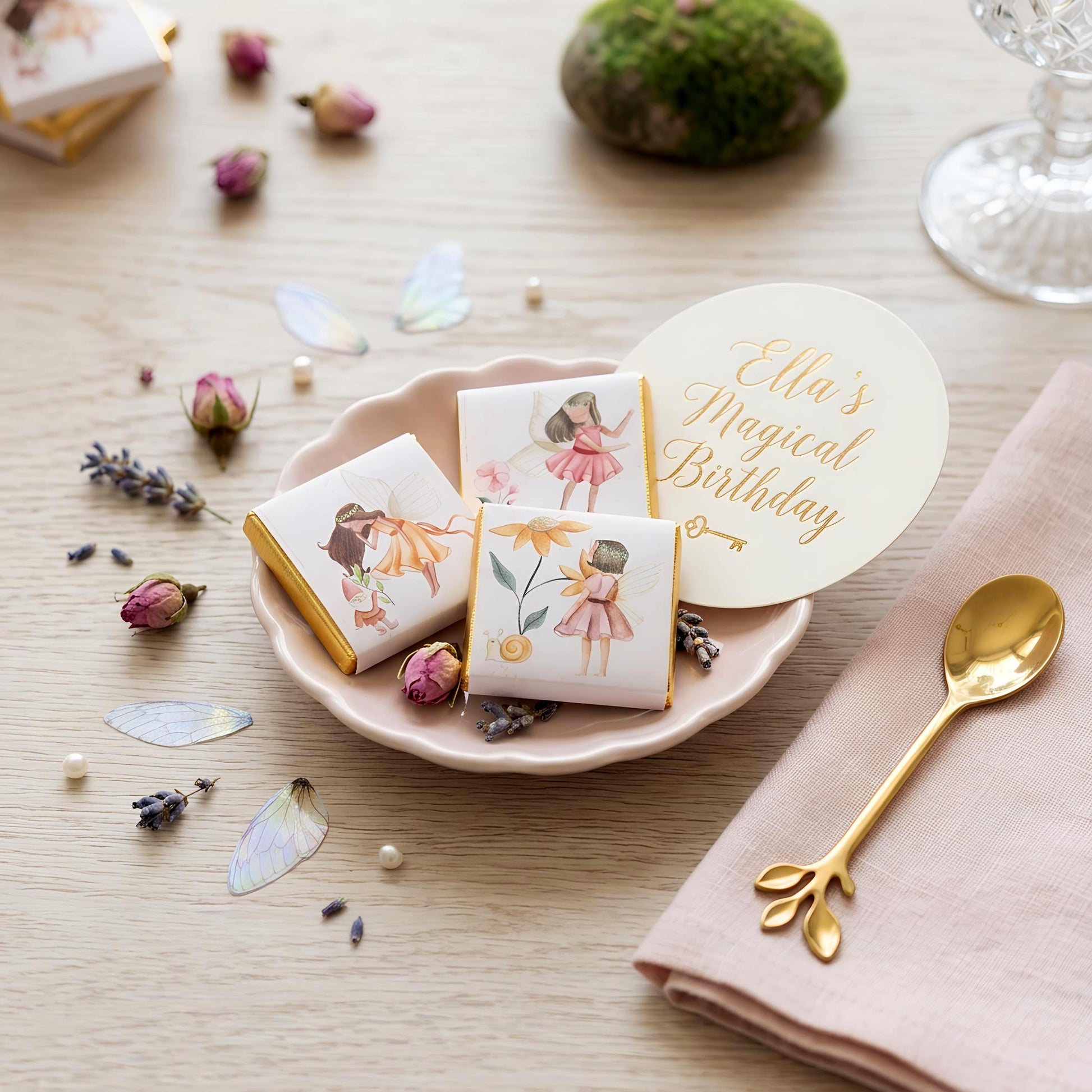 May include: Chocolates with fairy illustrations and gold foil edges, presented on a pink, scalloped plate. The scene includes a gold spoon and a card with the text "Ella's Magical Birthday". Dried flowers and fairy wings add to the whimsical theme.