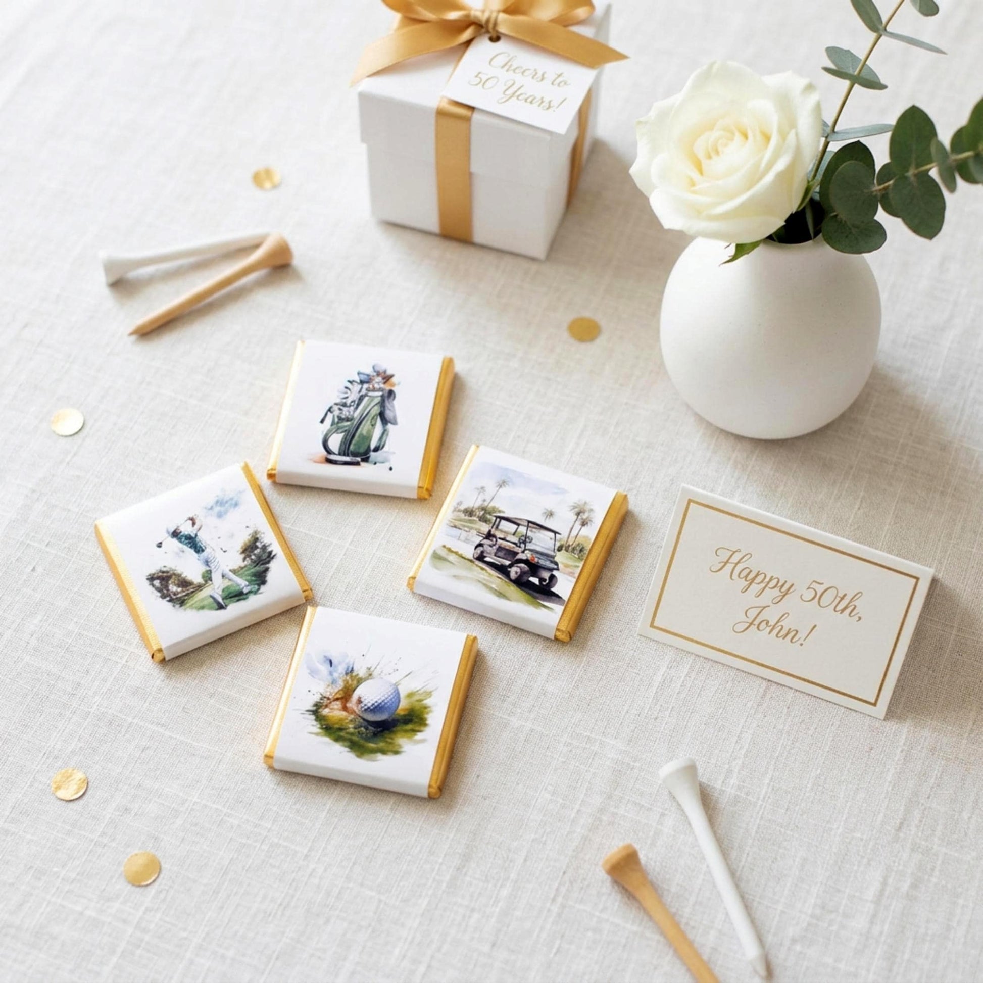 May include: Golf-themed party favors, including chocolates with golf course illustrations, a gift box with a gold ribbon, and a card that reads "Happy 50th, John!" A white rose in a small vase is also present.