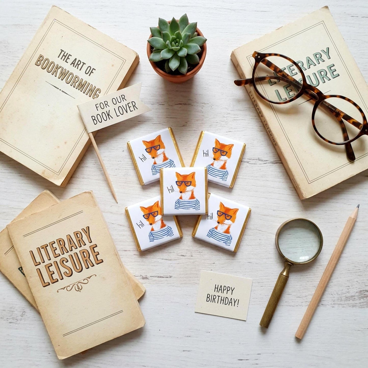 May include: Overhead view of vintage books, a succulent, and chocolates with a fox illustration. The chocolates feature a fox in glasses and a striped shirt. A sign reads "For Our Book Lover" and "Happy Birthday!"