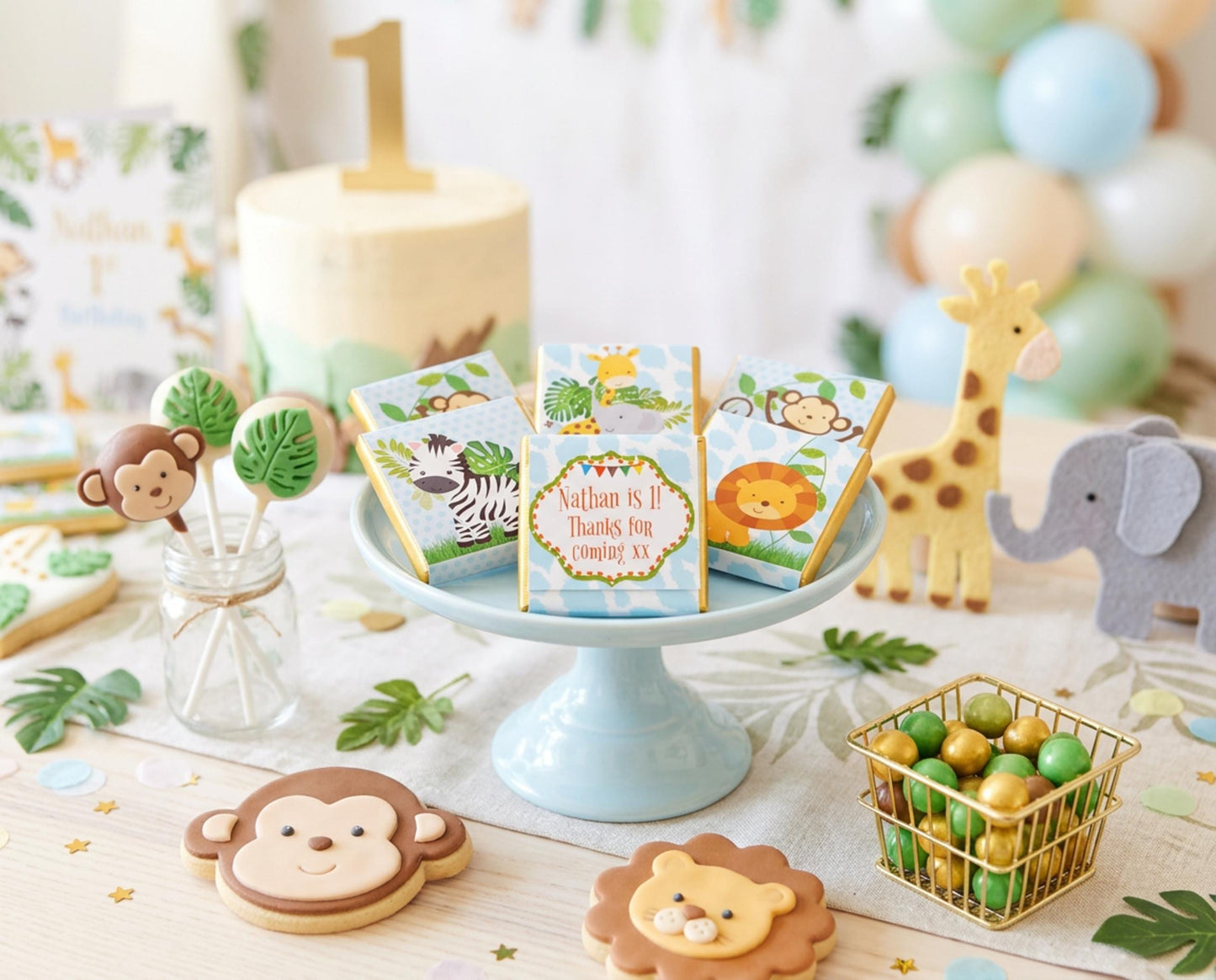 May include: A jungle-themed first birthday party scene. Features a cake with a gold number 1, animal-shaped biscuits, cake pops, and gift boxes. A giraffe and elephant toy are also present, along with balloons and a small wire basket filled with sweets.