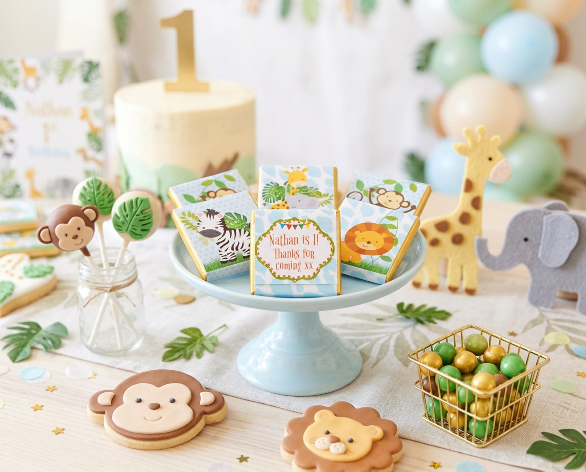 May include: A jungle-themed first birthday party scene. Features a cake with a gold number 1, animal-shaped biscuits, cake pops, and gift boxes. A giraffe and elephant toy are also present, along with balloons and a small wire basket filled with sweets.